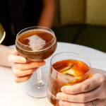 Two people seated at a table, clinking glasses filled with amber-colored drinks, each garnished differently, in a warmly lit environment.