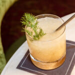 A cocktail with a sprig of dill and a metal straw, served on a napkin on a marble surface.