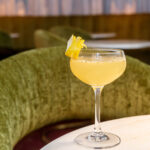 A cocktail with a lemon garnish on a marble table, with a plush green circular sofa in the background.