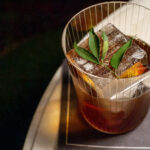 A glass of iced coffee with a metal wire cover, garnished with a bay leaf and a slice of dehydrated orange on a saucer.