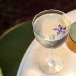 A clear stemmed cocktail glass with a floral garnish on a marble table, next to a gold lamp and a green velvet chair.